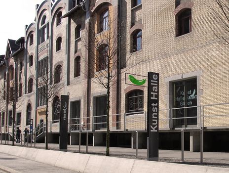 Das Bild zeigt ein Backsteingebäude. Davor steht eine grosse schwarze Tafel, die mit Kunst Halle angeschrieben ist. Aus der Tafel ragt auch ein Schild, das eine grüne Bratwurst zeigt. Das Schild bedeutet, dass das Museum geöffnet hat.