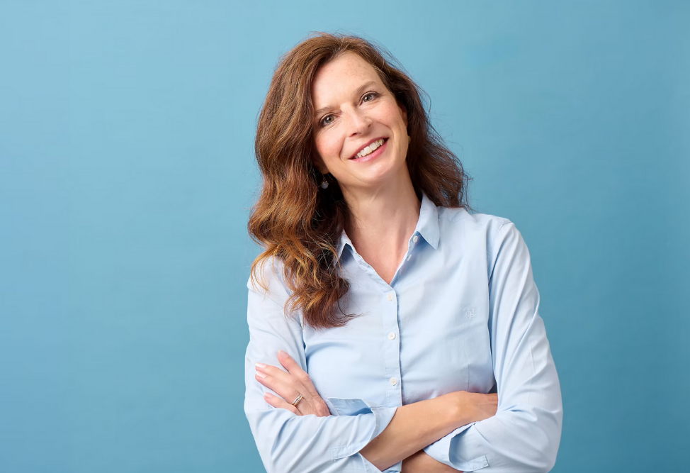 Das Bild ist ein Porträt von Sara Stocker vor blauem Hintergrund. Sara Stocker hat Inkluseum gegründet. Sie trägt eine helle Blus und hat langes braunes Haar. Sie hat die Arme vor der Brust verschränkt,d en Kopf leicht zur Seite geneigt und lächelt freundlich in die Kamera.
