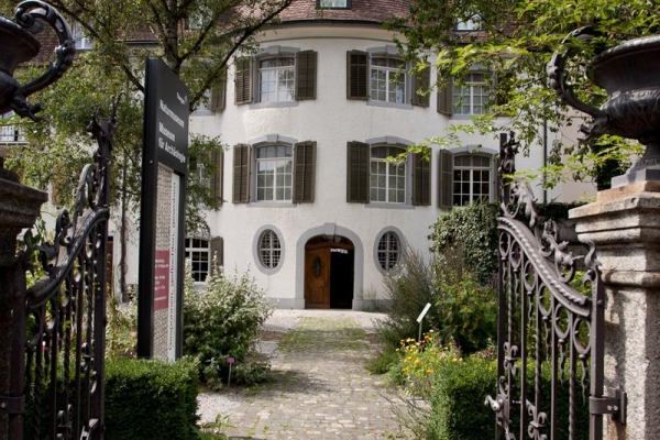 Auf dem Bild blickt man durch ein geöffnetes Eisentor auf ein weisses Gebäude. Ein Weg aus Pflastersteinen führt zum Eingang. Dieser ragt wie ein Halbrund hervor.