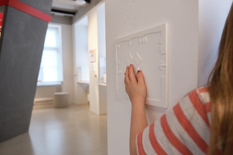 Das Bild zeigt ein Kind von hinten in einem Ausstellungsraum des Henry Dunant Museum. Das Kind ertastet mit der Hand einen Relief an der Wand. Das Relief macht den Raum für blinde Menschen fühlbar, damit sie sich orientieren können.