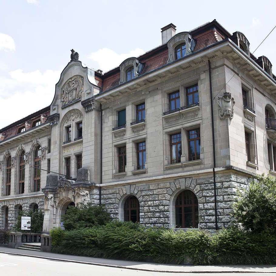 Das Bild zeigt ein altes Steingebäude mit drei Stockwerken an einer Strassenecke. Rund um das Gebäude wachsen grüne Hecken.