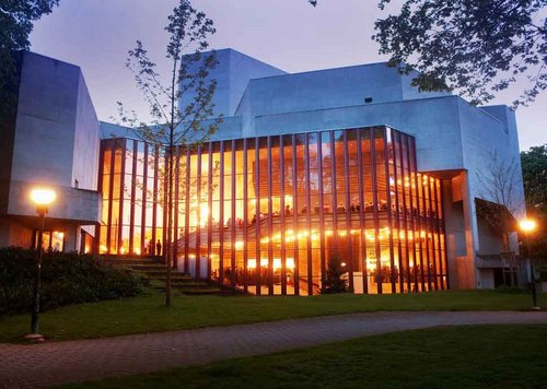 Das Bild zeigt das Theatergebäude. Es ist ein wüfelartiger Betonbau mit einem grossen Glasanbau. Auf dem Bild ist es Abend und das Gebäude leuchtet von innen. Weil das Theater in einem Park steht, hat es Bäume im Vordergrund.