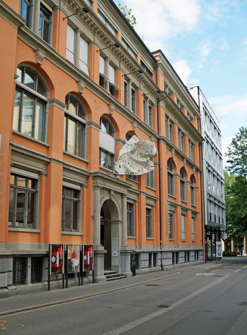 Das Bild zeigt ein rotes Steingebäude an einer ruhigen Strasse. Über dem Eingangstor hängt eine Skulptur aus Eisen, die aussieht wie ein Stück bestickter Stoff.