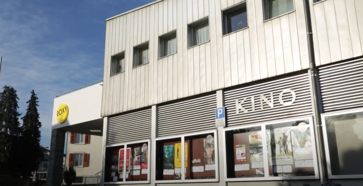 Das Bild zeigt ein modernes, eckiges Gebäude in grauer Farbe. An der Fassade hat es viele Schaukästen, in denen Filmplakate hängen, darüber steht in grossen Buchstaben Kino. Über dem Eingangsbereich an der Ecke vom Gebäude steht in schwarzer Schrift auf einem gelben Kreis der Name des Kinos: Roxy.